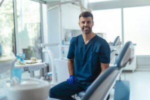 A smiling dentist in scrubs sitting in an operatory