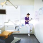 A dentist in scrubs sitting in an exam room looking at a tablet.