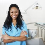 A woman smiling in dental scrubs by an exam chair